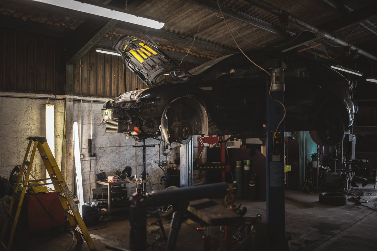 A vehicle is elevated on a hydraulic lift inside a workshop, with its hood open revealing engine components and tools scattered around