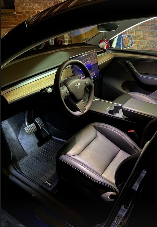Interior of a Tesla vehicle showing steering wheel, touchscreen display, light wood dashboard trim, and cream-colored leather seats with exposed door