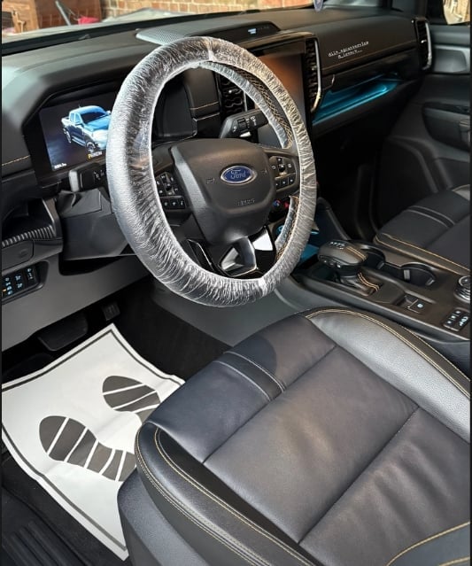 Car interior view showing Ford steering wheel with clear plastic protective cover, dark dashboard, and black leather seats with floor mats